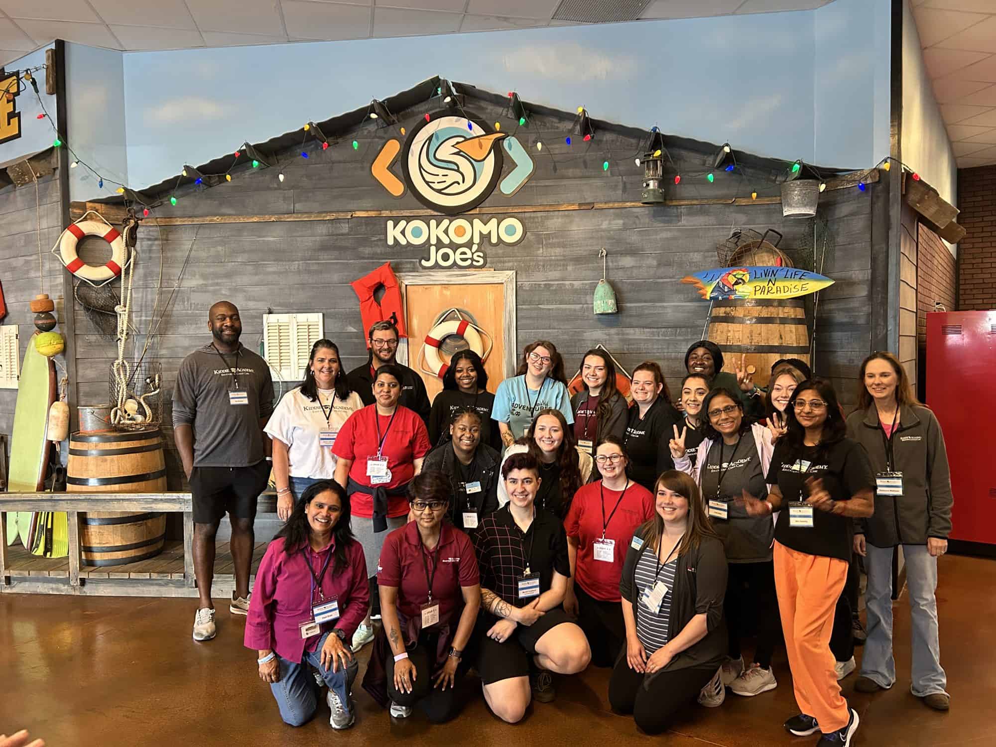 A smiling group poses for a photo in front of the Kokomo Joe's beach shack, decorated with string lights and the Kokomo Joe's logo